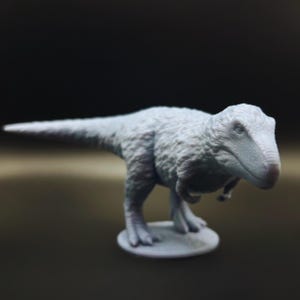 May include: A light gray 3D printed dinosaur figurine. The dinosaur is standing on a round base and has a textured surface.