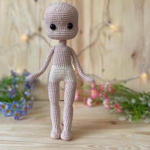 Puede incluir: Una muñeca de ganchillo con cuerpo rosa claro y parte inferior blanca. La muñeca tiene cabeza redondeada, ojos negros y brazos extendidos. La muñeca está sobre una superficie de madera.
