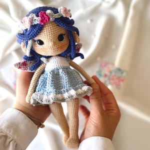 May include: A crocheted doll with blue hair, a flower crown, and a light blue dress. The doll has wings and is holding a small flower.
