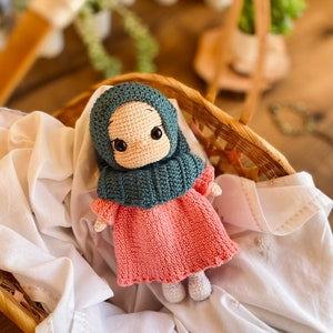 May include: A crocheted doll wearing a teal headscarf and a pink dress. The doll is sitting in a wicker basket lined with white fabric.