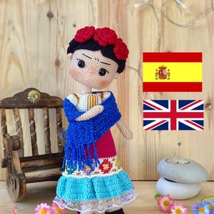 以下が含まれることがあります： 手作りのかぎ針編み人形。黒い髪に赤い花の冠、青いショール、カラフルなドレスを着ています。人形は詳細な顔をしており、木製の表面に立っています。装飾用の花が人形の周りに散らばっています。