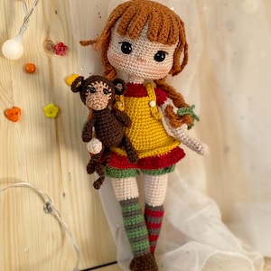 Puede incluir: Muñeca de ganchillo hecha a mano con pelo rojo y vestido amarillo, sosteniendo un pequeño mono marrón. La muñeca tiene una cara blanca con ojos negros y lleva calcetines a rayas rojas y verdes. La muñeca mide unos 25 cm de altura.