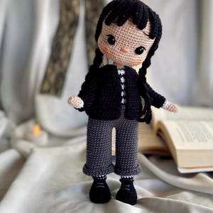 Peut inclure: Poupée au crochet faite à la main, avec des cheveux noirs tressés, vêtue d'une veste noire, d'un pantalon gris et de chaussures noires. La poupée a un visage beige et se tient sur une surface blanche. Article fait main.