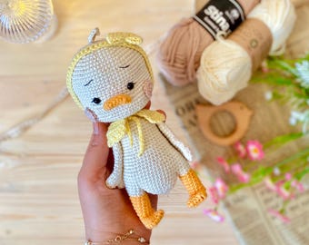 May include: A handmade crochet duck doll with a yellow bonnet, beak, and feet. The doll has black eyes and pink cheeks. The duck is made of light gray yarn and is a charming, soft toy.