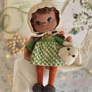 Può includere: Una bambola all'uncinetto con la pelle marrone, che indossa un vestito verde e bianco, un cappello marrone e scarpe marroni. La bambola tiene un coniglietto bianco all'uncinetto.