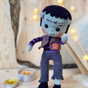 Pode incluir: Boneca Frankenstein de crochê feita à mão. A boneca tem um rosto verde, cabelo preto e veste uma roupa roxa e marrom. Os braços da boneca estão estendidos e ela usa sapatos pretos com detalhes brancos. O fundo inclui velas e um livro.