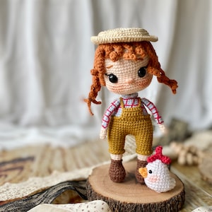 May include: A crocheted doll wearing a straw hat, overalls, and a plaid shirt, standing next to a crocheted white chick with a red comb. The doll and chick are on a wooden slice.