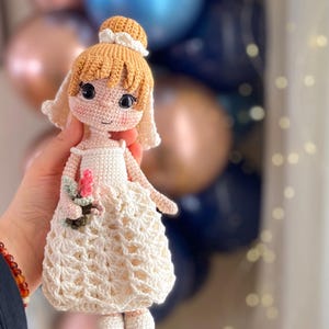 May include: A handmade crochet doll with a light skin tone, blonde hair, and a white dress. The doll is holding a small bouquet of pink flowers. The doll's dress has a detailed, openwork pattern. The background is blurred, with blue and gold balloons.