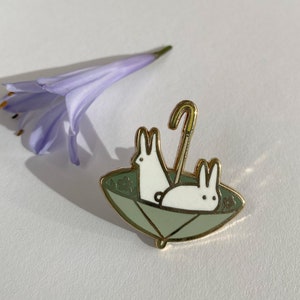 May include: Gold enamel pin featuring two white rabbits under a green umbrella with a gold handle.