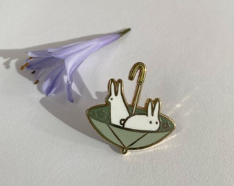 May include: Gold enamel pin featuring two white rabbits under a green umbrella with a gold handle.