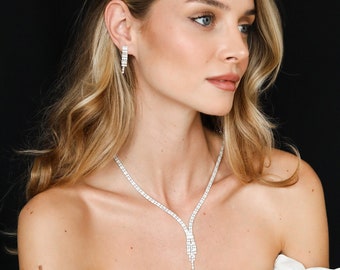 May include: A woman wearing a white dress with a sparkling tiara, necklace, and earrings. The tiara is a crown with a pointed top and is adorned with clear rhinestones. The necklace is a delicate chain with a pendant that has a teardrop shape and is also adorned with clear rhinestones. The earrings are long and dangle, also adorned with clear rhinestones.