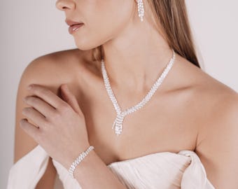 May include: A bride wearing a white strapless gown with a crystal necklace, bracelet, and earrings. She is wearing a crystal hairpiece.
