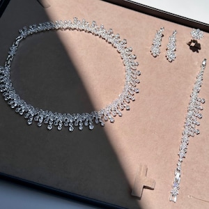 May include: A silver necklace, bracelet, and earrings set with clear crystal stones. The necklace has a teardrop design and the bracelet and earrings have a similar design.