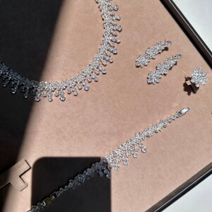 May include: A silver necklace, bracelet, and earrings set with clear crystal stones. The jewelry is displayed on a brown background.