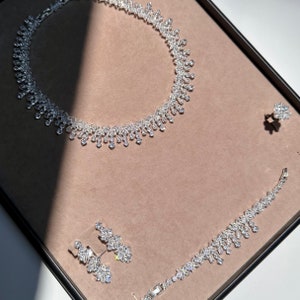May include: A silver necklace and bracelet set with sparkling clear stones. The necklace is a delicate chain with a pendant that features a cluster of stones. The bracelet is a simple chain with a matching pendant.