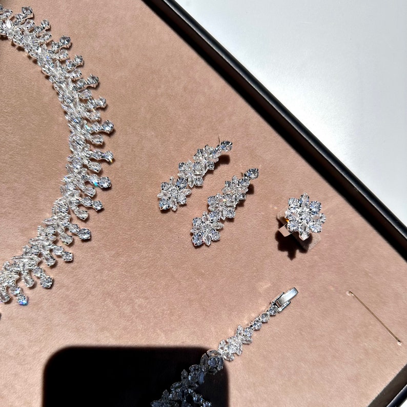 May include: A silver necklace, earrings, and ring set with clear cubic zirconia stones. The jewelry is arranged on a brown surface.