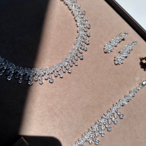 May include: A silver necklace and earrings set with a teardrop design. The jewelry is made of clear rhinestones.