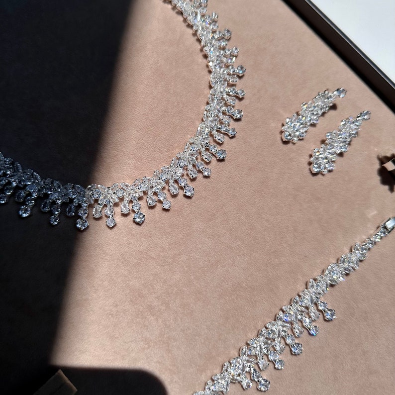 May include: A silver necklace and earrings set with a teardrop design. The jewelry is made of clear rhinestones.