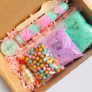 May include: A craft kit in a cardboard box containing colourful sensory materials. Includes bags of purple and green foam beads, a jar of gold glitter, and a clear tube with pink, green, and purple sand. A wooden spoon and a small jar of green beads are also included.