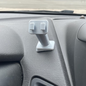 May include: A light gray, 3D-printed car phone mount. The mount is attached to a dark gray dashboard. The mount has a rectangular base, an angled arm, and a phone cradle with two raised tabs.