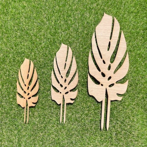 May include: Three wooden Monstera leaf cutouts in different sizes. The leaves feature detailed cut-out patterns and are light brown. Each leaf has a pointed stem. The leaves are displayed on a green surface.