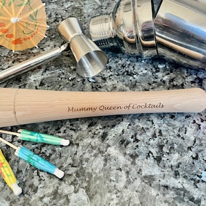 May include: A wooden cocktail muddler with the text "Mommy Queen of Cocktails" engraved on it. The muddler is lying on a grey countertop with a cocktail shaker, a jigger, and several colourful cocktail umbrellas.