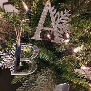 May include: Two silver glitter Christmas decorations shaped like the letters A and B, with a snowflake design on each decoration. The decorations are hanging on a green Christmas tree.