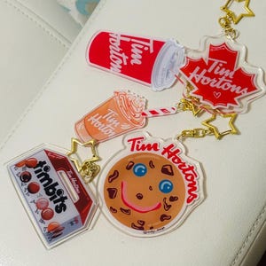 Puede incluir: Una colección de llaveros acrílicos con temática de Tim Hortons. Los llaveros presentan una taza de café roja, una hoja de arce, una galleta con una cara sonriente, una caja de Timbits y una bebida de café. Cada llavero está unido a una cadena dorada.
