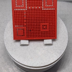 Puede incluir: Una plantilla de plástico roja con un patrón de cuadrícula y tres recortes cuadrados. La plantilla está sobre una superficie blanca.