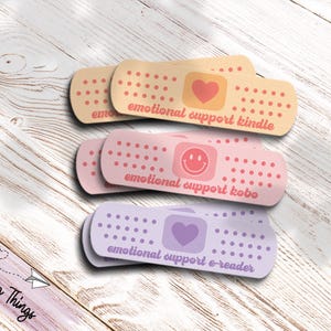 Könnte beinhalten: Drei Pflaster-förmige Aufkleber in Beige, Rosa und Lavendel. Jeder Aufkleber trägt den Text "emotional support" gefolgt von "kindle", "kobo" oder "e-reader" sowie ein Herz oder Smiley. Sie liegen auf einer hellen Holzoberfläche.