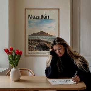 May include: A woman sits at a wooden table, drawing on a white sheet of paper with a black pen. A framed poster with the text "Mazatl&aacute;n" and a picture of a beach scene hangs on the wall behind her. A vase with red tulips sits on the table in front of her.