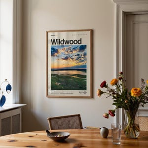 May include: A framed poster with the word "Wildwood" at the top, featuring a scenic landscape of a sunset over a body of water and marshland. The poster is displayed on a beige wall above a wooden table.