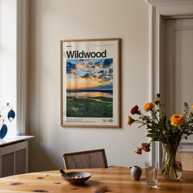 May include: A framed poster with the word "Wildwood" at the top, featuring a scenic landscape of a sunset over a body of water and marshland. The poster is displayed on a beige wall above a wooden table.