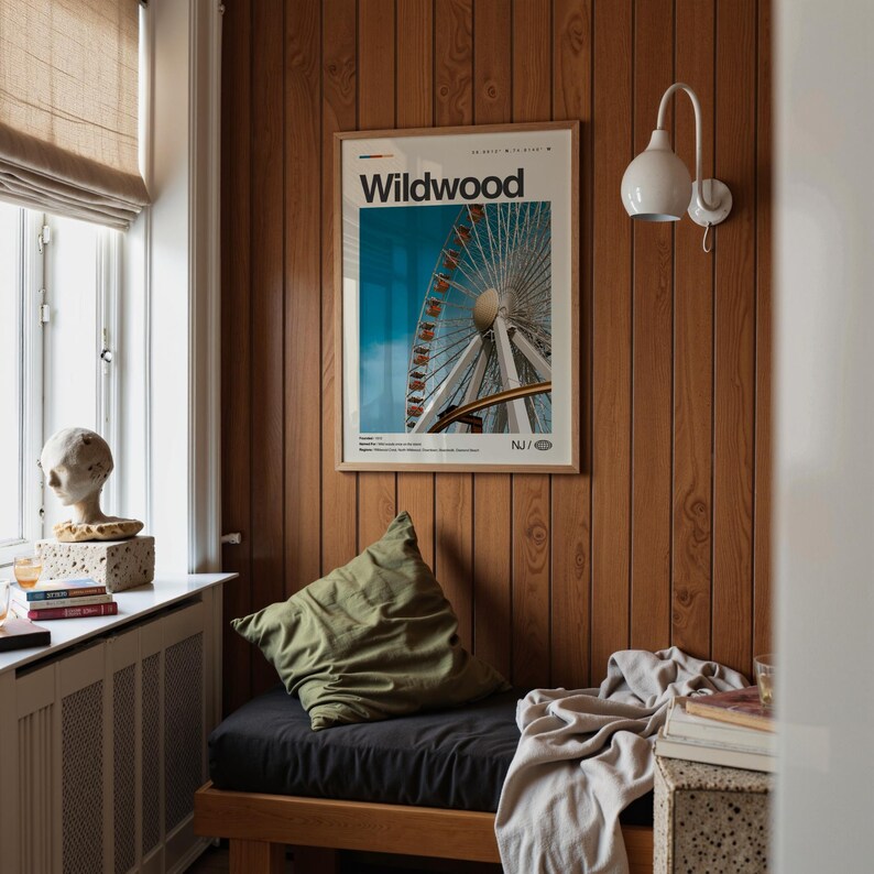 May include: A room with wood-paneled walls and a framed poster that reads "Wildwood" with a Ferris wheel image. A green pillow rests on a black cushion, and a white lamp is mounted on the wall. A window with a blind is visible.