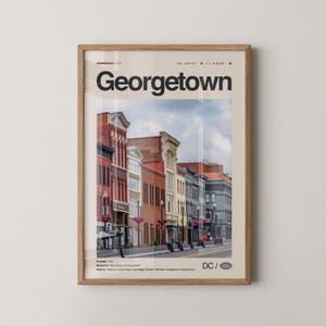 May include: A framed print featuring a street scene in Georgetown, DC. The print showcases a row of historic buildings in shades of red, beige, and gray. The word "Georgetown" is prominently displayed at the top, with the coordinates and "DC /" below.