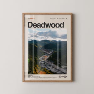 May include: A framed poster featuring the word "Deadwood" in bold black letters. The poster displays a scenic view of a town nestled in a valley surrounded by green hills and a cloudy sky. The poster has a light wood frame.
