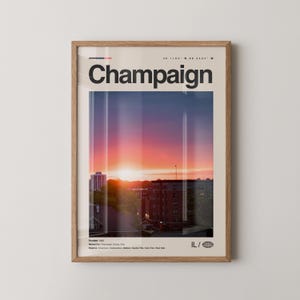 May include: Framed art print featuring the word "Champaign" in bold black letters above a colorful sunset cityscape. The print includes the coordinates 40.1164° N, 88.2434° W and the abbreviation "IL/". The print is in a light brown wooden frame.