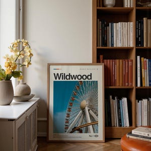 May include: A framed poster with the word "Wildwood" in bold, black letters. The poster features a Ferris wheel against a blue sky. The image is in a light brown wooden frame. A vase of yellow flowers sits on a white surface.