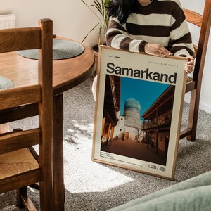May include: A framed poster with the word "Samarkand" in a bold font. The poster features a photograph of a building with a blue dome and wooden balconies. The poster is held by a person wearing a brown and white striped sweater.