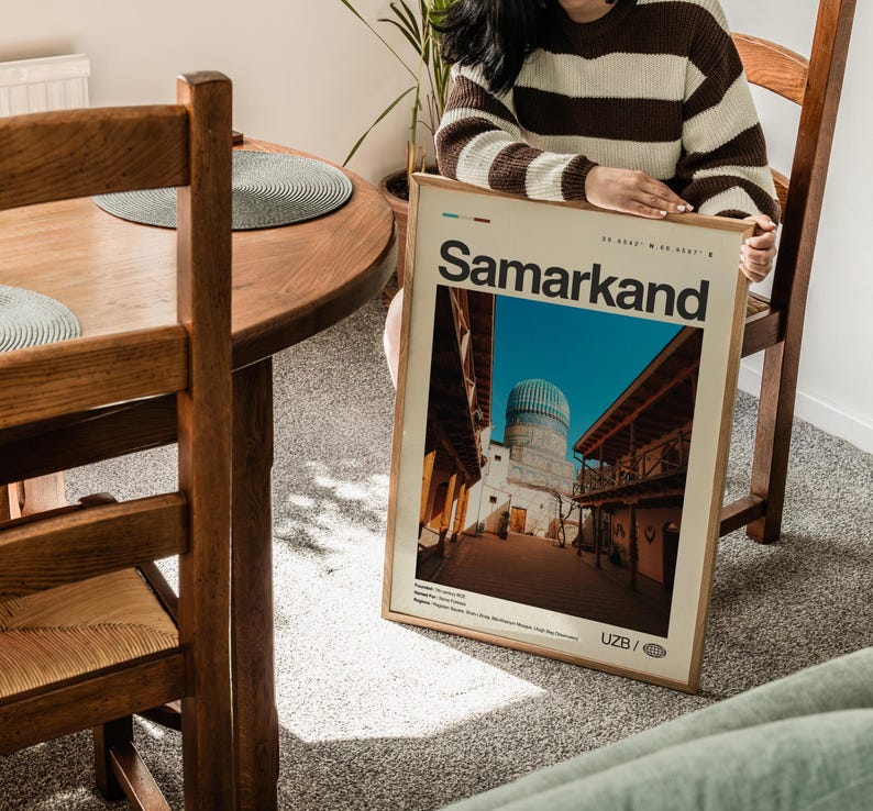 May include: A framed poster with the word "Samarkand" in a bold font. The poster features a photograph of a building with a blue dome and wooden balconies. The poster is held by a person wearing a brown and white striped sweater.