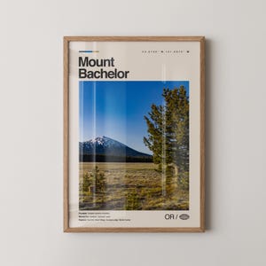 Può includere: Una stampa incorniciata con il Monte Bachelor, con una vetta innevata sotto un cielo blu. La stampa include il testo "Mount Bachelor" e le coordinate, con un paesaggio di alberi e campi. La cornice è di colore legno chiaro.
