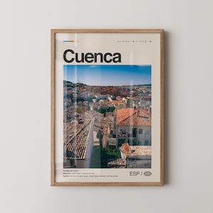 May include: A framed print featuring the word "Cuenca" in large black letters above a cityscape. The image shows a view of a city with red-tiled roofs and a blue sky. The print includes coordinates and the letters "ESP".