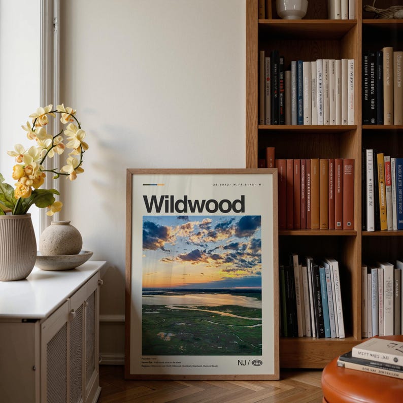 May include: A framed poster with the word "Wildwood" in bold black letters. The poster features a scenic landscape of a sunset over a body of water and marshland. The poster is displayed in a room with a bookshelf and decorative vases.