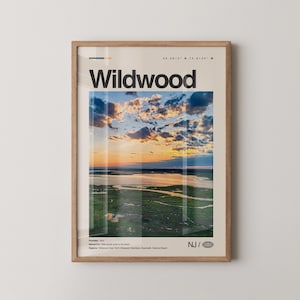 May include: A framed print featuring the word "Wildwood" in bold black letters. The image within the frame showcases a scenic aerial view of a coastal landscape at sunset, with a vibrant sky and green marshland. The print includes text indicating the location's coordinates and founding year.