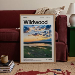 May include: A framed Wildwood print featuring a scenic aerial view of a coastal landscape at sunset. The print has a wooden frame and includes the text "Wildwood" in bold black letters. The image captures a vibrant sky with clouds and a body of water.