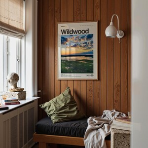 May include: A framed "Wildwood" poster with a landscape image hangs on a wood-paneled wall. Below, a dark cushion and olive green pillow sit on a wooden bench. A white wall lamp and a window with a beige shade are also visible.