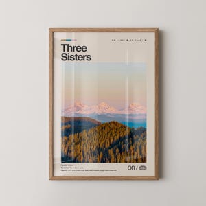Puede incluir: Un estampado enmarcado con un paisaje de las montañas Three Sisters en Oregón. El estampado tiene un fondo beige claro con las palabras "Three Sisters" en negro. La imagen muestra montañas cubiertas de nieve, un bosque y una masa de agua.