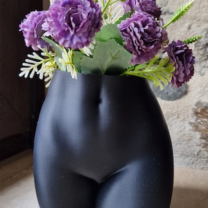 Può includere: Un vaso in ceramica nero a forma di torso umano, con fiori viola all'interno.