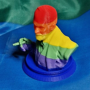 Op de afbeelding: Een 3D-geprint buste van een persoon met een rode hoed en een regenboogkleurige sjaal. Het buste staat op een blauwe basis.