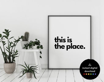 May include: Black and white printable wall art with the text "this is the place."  The print is framed in a black frame and is displayed on a white wall with plants.